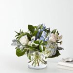 A lush floral arrangement in a clear cylindrical glass vase featuring white roses, light blue hydrangeas, blue delphiniums, and silvery dusty miller foliage against a clean white background.