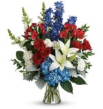 A patriotic-themed floral arrangement in a glass vase featuring red roses, white lilies, blue hydrangeas, blue delphinium, and red alstroemeria accented with dusty miller and greenery.