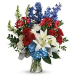 A vibrant patriotic-themed flower arrangement in a clear glass vase, featuring red roses, blue hydrangeas, white lilies, blue delphiniums, and white snapdragons.