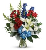 A vibrant floral arrangement in a glass vase featuring red roses, blue hydrangeas, white lilies, and red alstroemeria with green foliage.