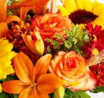 A vibrant autumn floral bouquet featuring orange lilies, bi-color orange and yellow roses, a large yellow sunflower, and red chrysanthemums accented with green filler and dried wheat.