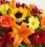 A vibrant autumnal floral arrangement featuring a prominent orange lily, a yellow sunflower with a dark center, orange roses, red daisies, and yellow chrysanthemums, accented with dried autumn leaves and green filler.