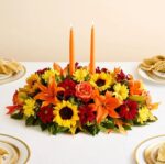 A horizontal fall floral centerpiece featuring sunflowers, orange lilies, red daisies, and roses, topped with two tall orange taper candles on a white tablecloth.