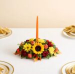 A horizontal autumn floral centerpiece featuring a large sunflower, yellow daisies, orange carnations, and dark red chrysanthemums, with a single tall orange taper candle standing in the center on a white tablecloth.