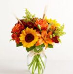 A vibrant autumn floral arrangement in a glass vase featuring sunflowers, orange lilies, roses, and red chrysanthemums accented with dried wheat stalks and green solidago.
