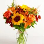 A vibrant autumn flower arrangement in a clear glass vase, featuring large yellow sunflowers, orange lilies, orange roses, and red chrysanthemums accented with dried wheat stalks and green foliage.