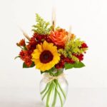 A vibrant autumn floral arrangement in a clear glass vase featuring a large sunflower, orange roses, bronze chrysanthemums, yellow solidago, and dried wheat stalks.