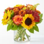 A vibrant autumn-themed floral arrangement in a clear glass cylinder vase, featuring sunflowers, orange roses, and burgundy chrysanthemums.