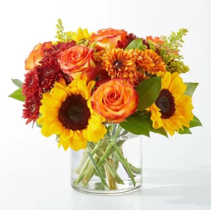A vibrant autumn-themed floral arrangement in a clear glass cylindrical vase, featuring bright yellow sunflowers, orange-tipped roses, deep red chrysanthemums, and green foliage against a white background.