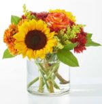 A vibrant autumn-themed floral arrangement featuring a large yellow sunflower, an orange and yellow bi-color rose, and deep red dahlias in a clear glass cylinder vase filled with water.