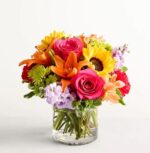 A vibrant and lush "Floral Embrace" flower arrangement featuring hot pink roses, bright yellow sunflowers, orange lilies, and purple stock flowers in a clear glass cylinder vase.