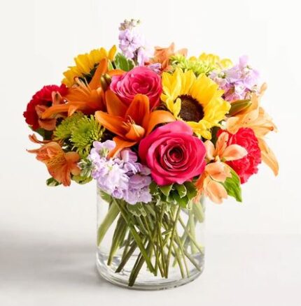 A vibrant and colorful mixed floral arrangement in a clear cylindrical glass vase, featuring bright sunflowers, hot pink roses, orange lilies, lavender stock flowers, and green chrysanthemums.