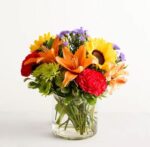 A vibrant floral arrangement in a clear cylindrical glass vase featuring orange lilies, a bright yellow sunflower, red carnations, purple asters, and green chrysanthemums.