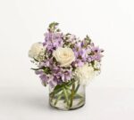 A beautiful floral arrangement in a clear glass cylinder vase featuring white roses, lavender alstroemeria, purple stock flowers, and delicate white baby's breath against a plain white background.