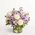A lush floral arrangement in a glass cylinder vase featuring white roses, lavender stocks, purple alstroemeria, and white baby's breath accented with green foliage.