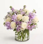 A premium floral arrangement featuring a mix of white roses, lavender stocks, purple alstroemeria, and white carnations in a clear cylindrical glass vase against a white background.