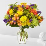 A vibrant "Marmalade Skies" floral bouquet in a clear glass vase featuring orange roses, yellow chrysanthemums, peach alstroemeria, purple statice, and green spider mums.