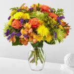 A vibrant "Marmalade Skies" flower bouquet featuring orange roses, yellow chrysanthemums, purple statice, and green spider mums in a clear glass vase.