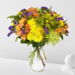 A vibrant "Marmalade Skies" flower bouquet featuring yellow chrysanthemums, orange alstroemeria, purple statice, and green spider mums in a clear glass vase on a white surface.
