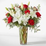 A lush floral arrangement in a clear glass cylinder vase featuring large white lilies, vibrant red roses, white stock flowers, and festive greenery with red berries.