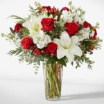 A festive holiday flower arrangement in a clear glass vase featuring red roses, white lilies, white stock flowers, red hypericum berries, and seasonal evergreen foliage.
