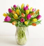 A vibrant bouquet of colorful tulips including pink, yellow, and orange varieties, arranged in a clear fluted glass vase against a plain white background.