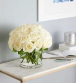 A dense bouquet of creamy white roses in a clear glass cylindrical vase, placed on a white marble console table with gold trim, set against a light grey wall.