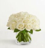 A lush bouquet of creamy white roses tightly arranged in a low, clear glass cylindrical vase, featuring vibrant green leaves and visible stems through the water.
