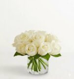 A modern arrangement of fresh white roses in a low, clear glass cylindrical vase against a minimalist white background