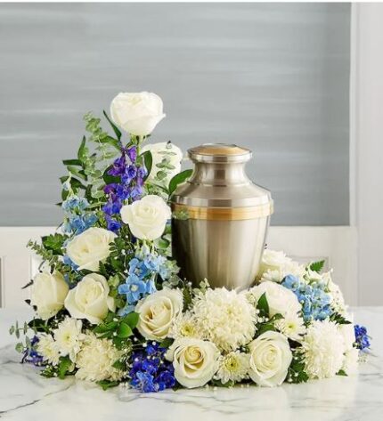 A premium cremation memorial wreath featuring white roses, white chrysanthemums, and blue delphinium surrounding a brushed metal urn with gold accents.