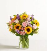 A vibrant and lush floral arrangement in a clear cylindrical glass vase, featuring bright yellow sunflowers, soft pink roses, purple stock flowers, pink alstroemeria, and green filler foliage against a white background.