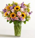 A large, vibrant "Garden of Grandeur" floral bouquet in a clear glass vase, featuring yellow sunflowers, light pink roses, purple stock flowers, and green accents.