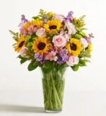 A large, vibrant "Garden of Grandeur" premium floral bouquet in a clear glass vase, featuring yellow sunflowers, light pink roses, purple stock flowers, pink alstroemeria, and green filler accents.