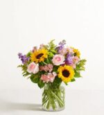 A vibrant floral arrangement in a clear glass cylinder vase featuring sunflowers, pink roses, purple stock, pink alstroemeria, and green accent fillers against a plain white background.