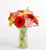 A vibrant bouquet of colorful Gerbera daisies in shades of red, pink, orange, and yellow, arranged in a clear fluted glass vase against a plain white background.