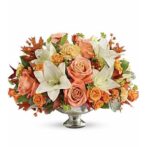 A lush autumn floral centerpiece featuring peach roses, white lilies, and orange spray roses in a silver pedestal bowl.