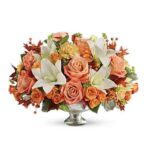 A lush autumn floral centerpiece featuring peach roses, white lilies, orange spray roses, and yellow carnations arranged in a silver mercury glass pedestal bowl.