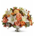 A lush autumn floral arrangement featuring white lilies, orange roses, and peach carnations in a silver mercury glass pedestal bowl.