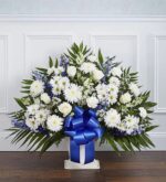 A large, fan-shaped funeral floor basket arrangement featuring white roses, chrysanthemums, and carnations accented with blue delphinium and lush greenery, finished with a prominent blue satin ribbon.