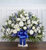 A large, fan-shaped funeral floral arrangement featuring white roses, carnations, daisies, and chrysanthemums mixed with blue delphinium and accented by a prominent blue satin ribbon in a white floor basket.
