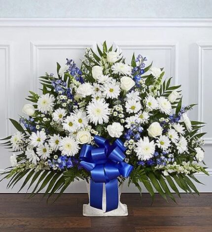 A large, fan-shaped funeral floral arrangement featuring white roses, carnations, daisies, and chrysanthemums mixed with blue delphinium and accented by a prominent blue satin ribbon in a white floor basket.