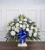 A large, fan-shaped funeral flower arrangement in a white floor basket, featuring white roses, daisies, and carnations accented with blue delphinium and a large blue satin bow.
