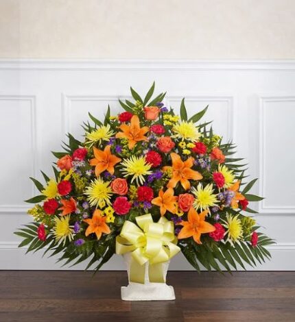 A large, fan-shaped funeral floor basket arrangement featuring vibrant orange lilies, red carnations, orange roses, and yellow spider mums, accented with purple statice and lush greenery, finished with a large yellow satin bow in a white pedestal container.