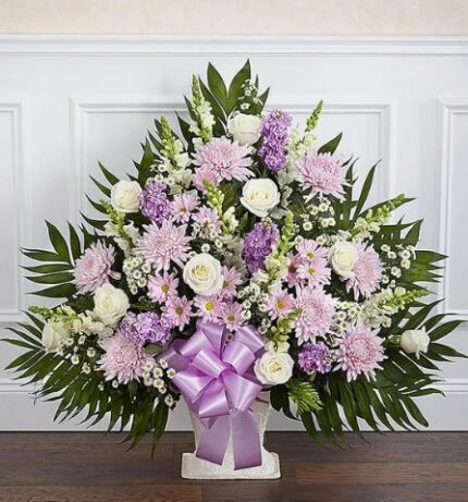 A large, fan-shaped funeral floor basket arrangement featuring a mix of lavender and white flowers, including roses, chrysanthemums, and snapdragons, accented with lush greenery and a purple satin bow.
