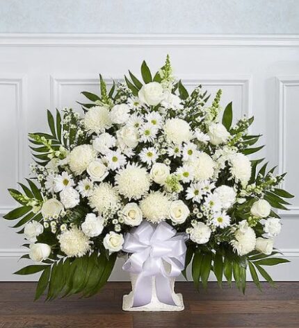 A large, fan-shaped funeral floor basket arrangement featuring a variety of premium white flowers including roses, lilies, chrysanthemums, and carnations, accented with lush greenery and a white satin ribbon.