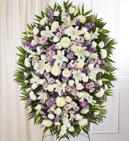 A large, oval-shaped funeral standing spray featuring a dense arrangement of white lilies, roses, chrysanthemums, and lavender-colored daisies and stock flowers, accented with lush green palm fronds and emerald foliage.