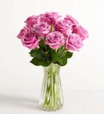 A dozen premium long-stemmed lavender-purple roses arranged in a clear fluted glass vase against a minimalist white background.