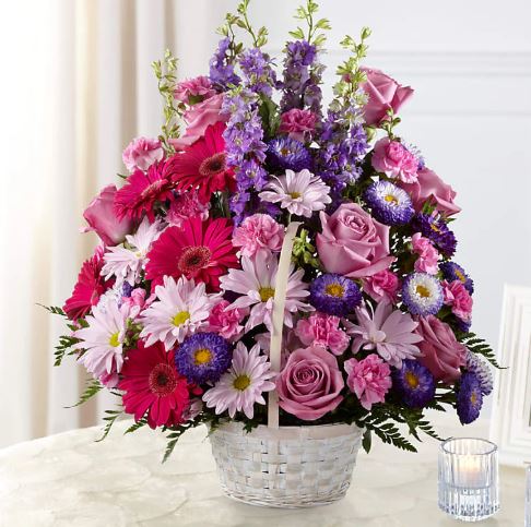 A large, vibrant floral arrangement in a white wicker basket featuring pink gerbera daisies, purple roses, lavender larkspur, and purple asters set against a soft, light background.