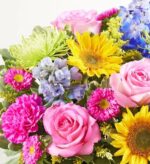 A vibrant and colorful floral bouquet featuring pink roses, bright yellow sunflowers, green spider mums, and purple delphiniums.