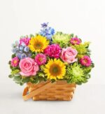 A vibrant and colorful floral arrangement featuring sunflowers, pink roses, magenta carnations, and green chrysanthemums tightly packed in a rustic woven wooden basket with a handle.